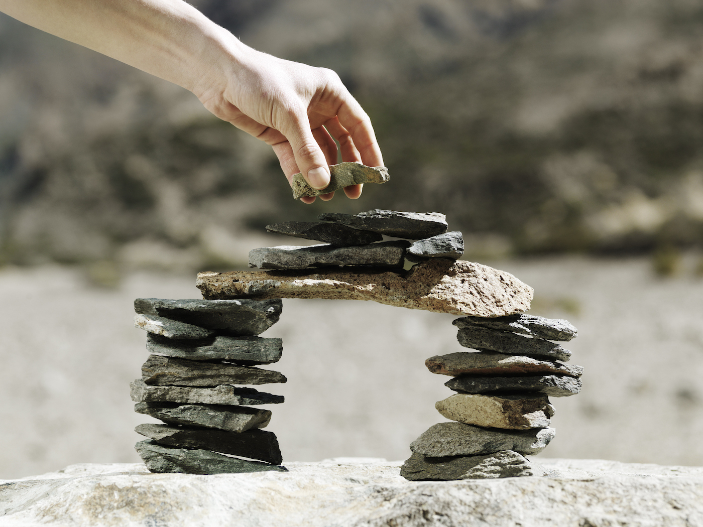 Final stone being placed by hand on a balancing miniature model bridge made of small flat rocks outside